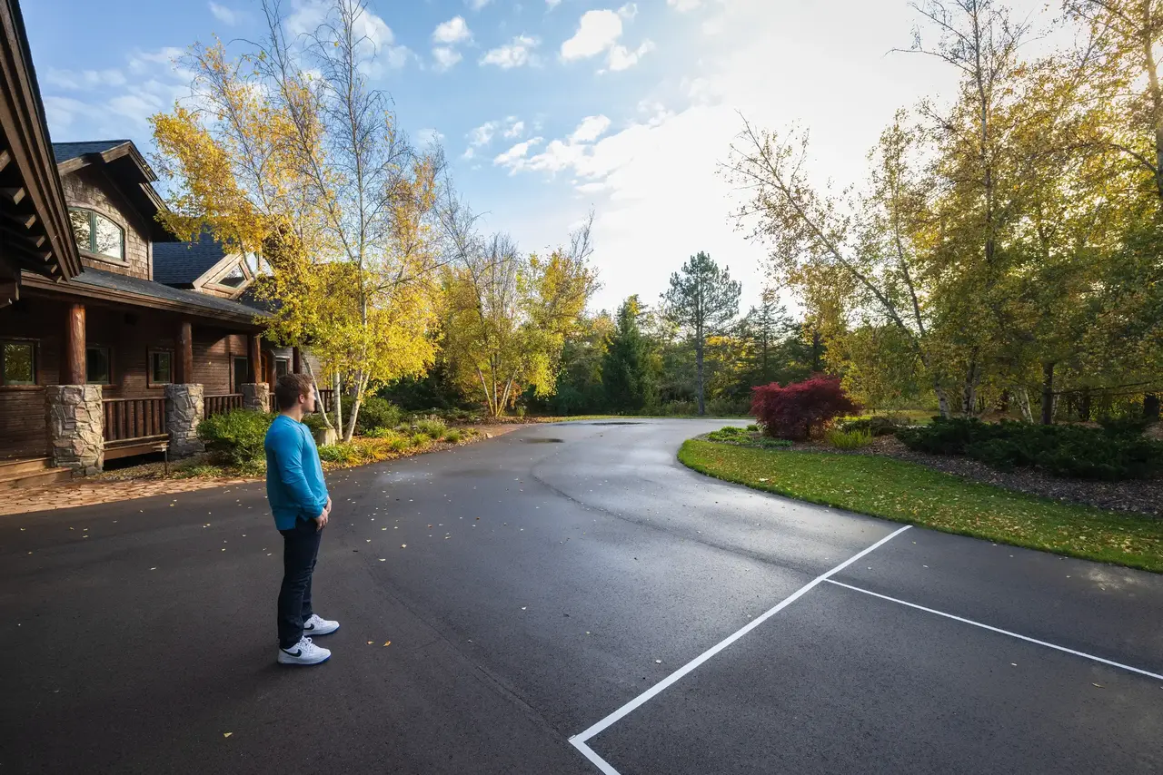 A person stands on a paved driveway, looking towards trees with autumn foliage. A large wooden house is on the left, and the sky is partly cloudy.