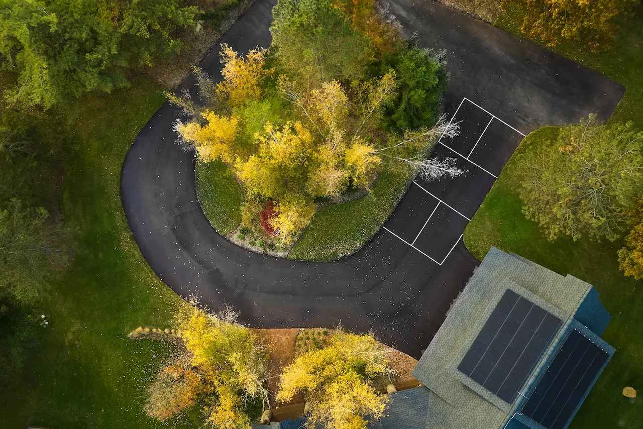 Trees with yellow and green foliage stand in the center of a circular driveway. Solar panels are visible on a nearby roof, set in a grassy, landscaped area.